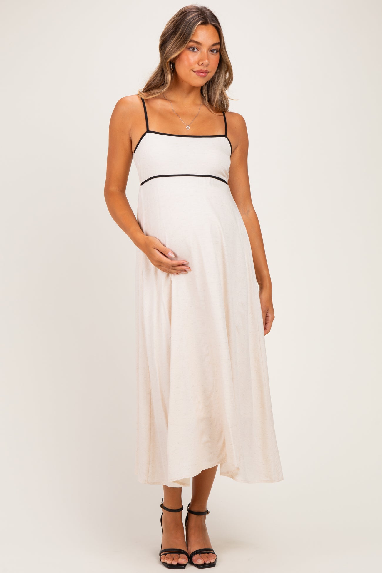woman wearing a beige maternity dress with black trim, posing with her baby bump visible