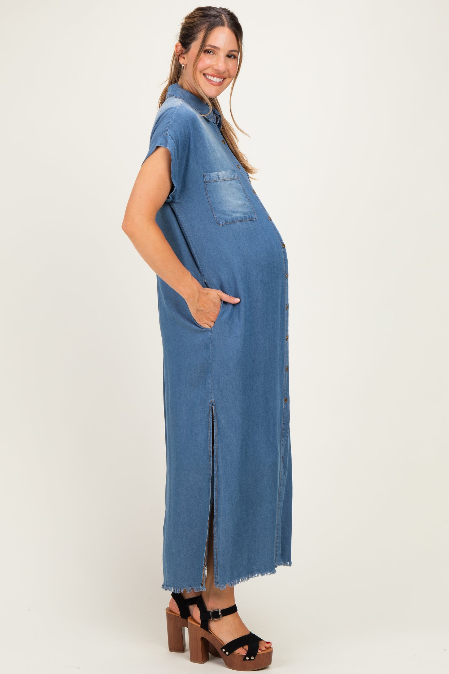woman wearing a blue denim maternity dress with short sleeves, side pockets, and a front slit, showcasing her baby bump