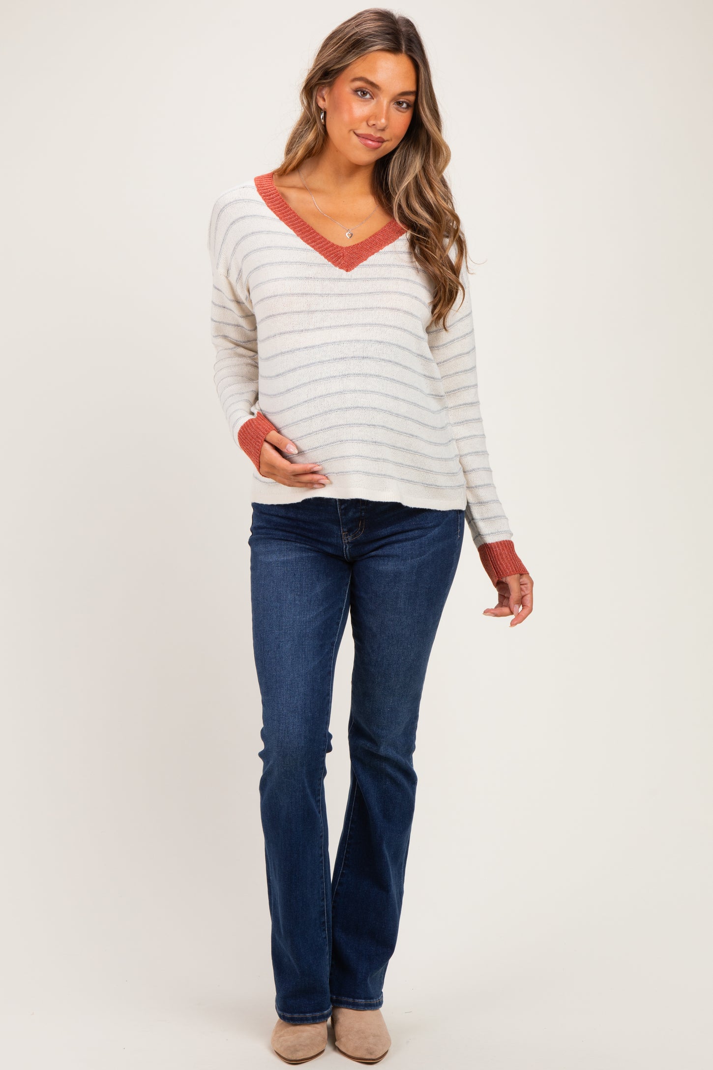 woman wearing a cream and rust striped maternity sweater with a v-neck and blue maternity jeans, holding her baby bump