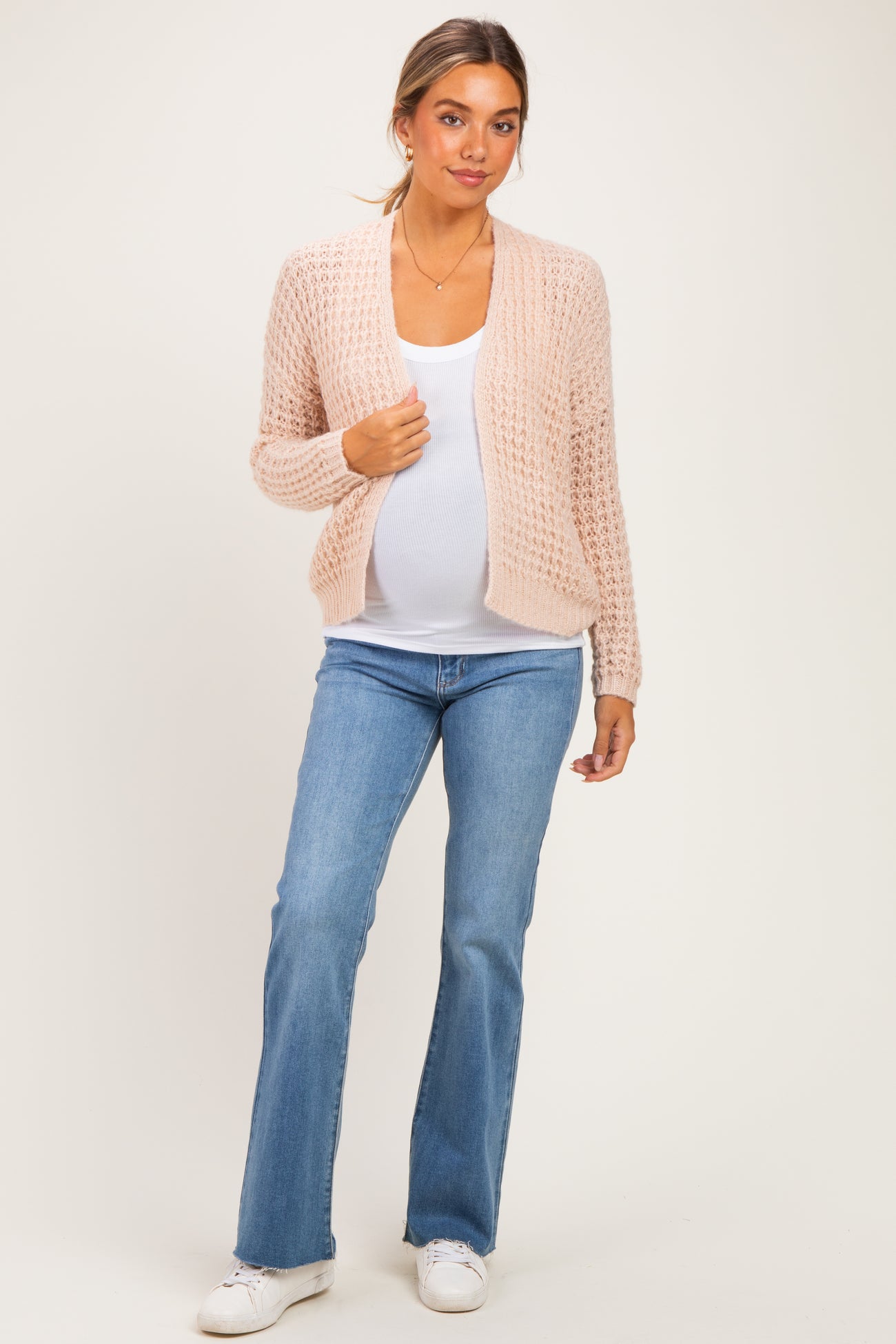 woman wearing a light pink open knit maternity cardigan over a white tank top and blue jeans, showcasing her baby bump