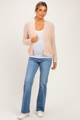 woman wearing a light pink open knit maternity cardigan over a white tank top and blue jeans, showcasing her baby bump