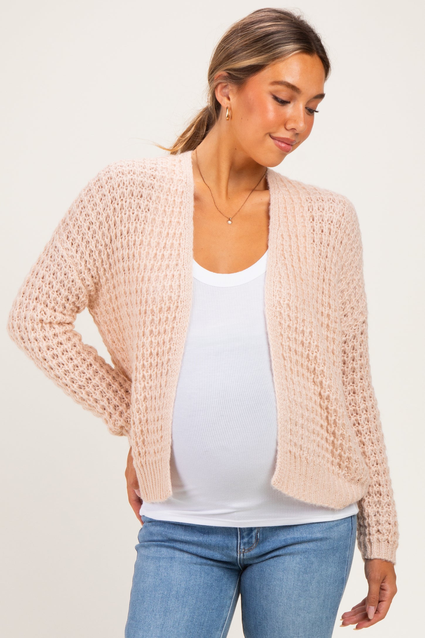 woman wearing a light pink open-knit maternity cardigan over a white tank top, showcasing her baby bump