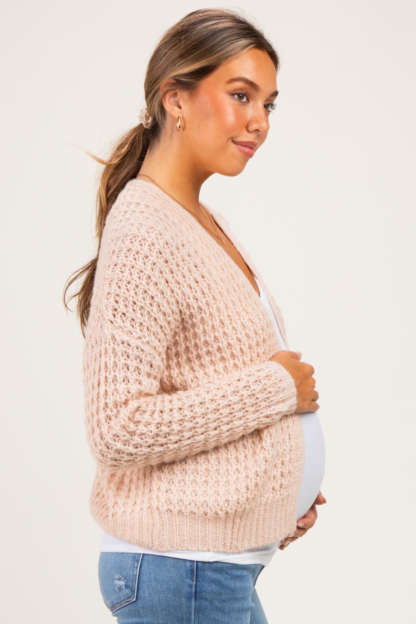 woman wearing a light pink maternity cardigan and holding her baby bump while facing to the side