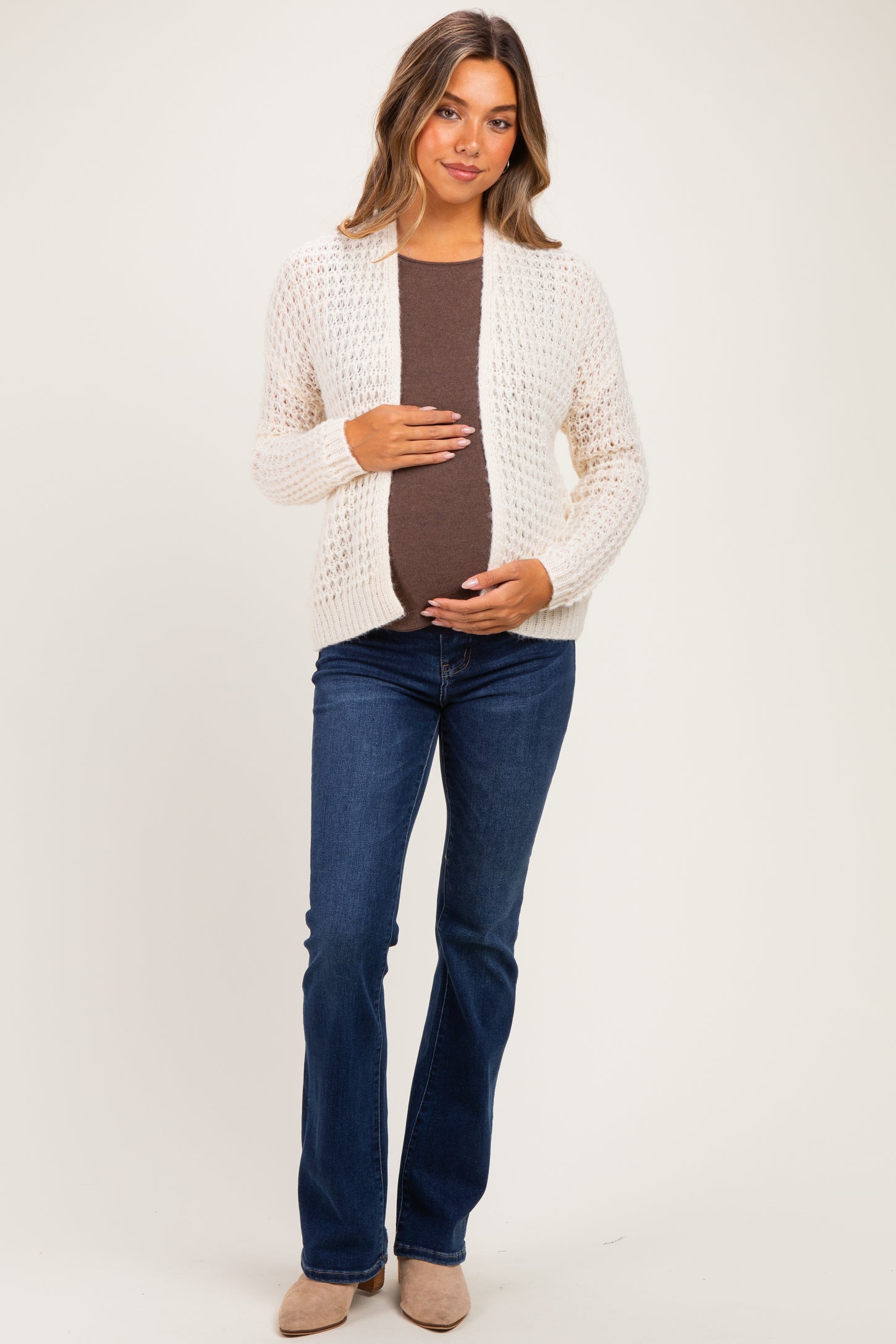 woman wearing a cream open-knit maternity cardigan over a brown tank top, cradling her baby bump, paired with blue flared jeans
