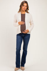 woman wearing a cream open-knit maternity cardigan over a brown tank top, cradling her baby bump, paired with blue flared jeans