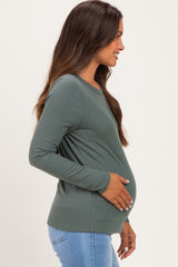 woman wearing a dark green long sleeve maternity top, gently holding her baby bump while smiling