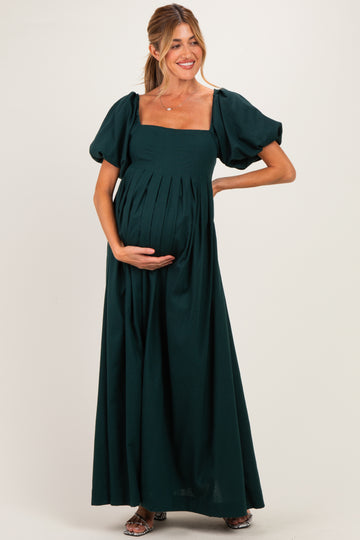 woman wearing a dark green maternity maxi dress with puff sleeves and a pleated bodice, holding her baby bump