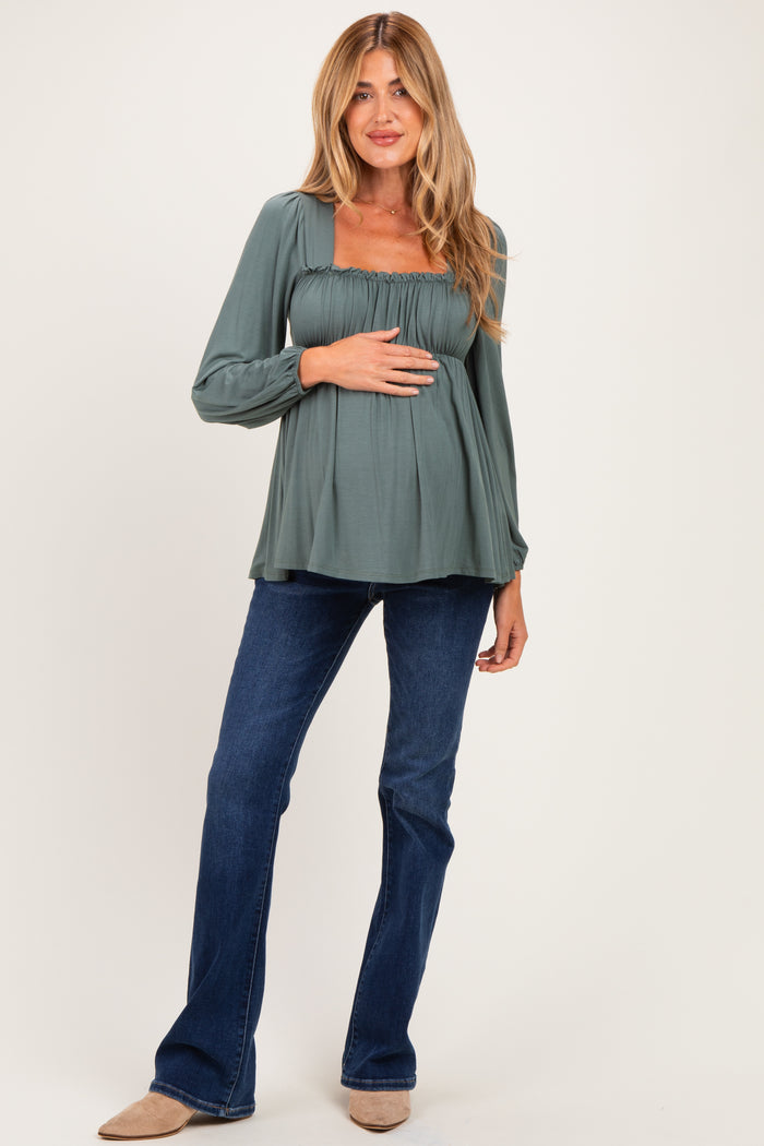 woman wearing a sage green long sleeve maternity top with smocked detailing and a visible baby bump, paired with flared jeans