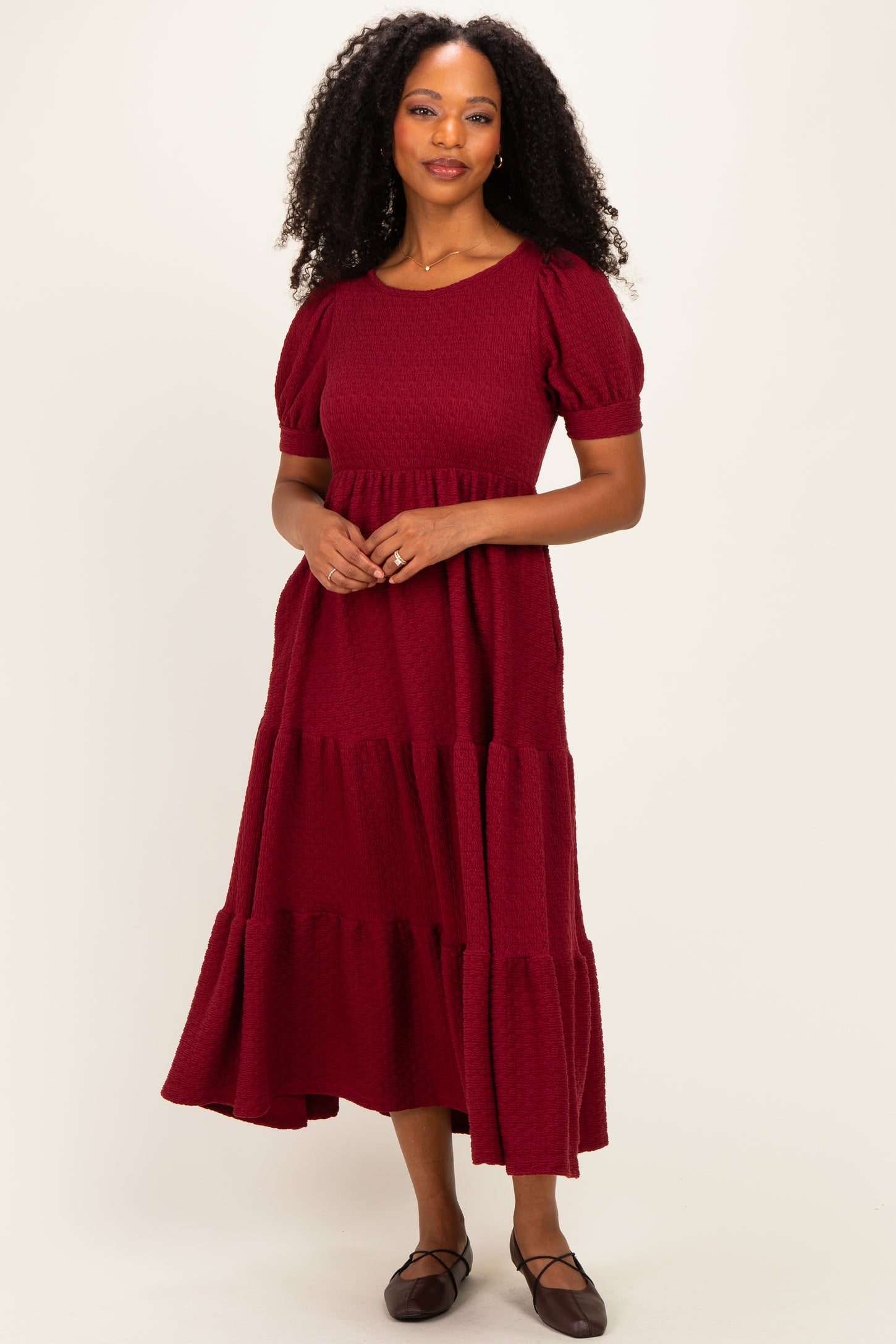 woman wearing a burgundy tiered maxi dress with puff sleeves and a textured fabric, standing against a plain background