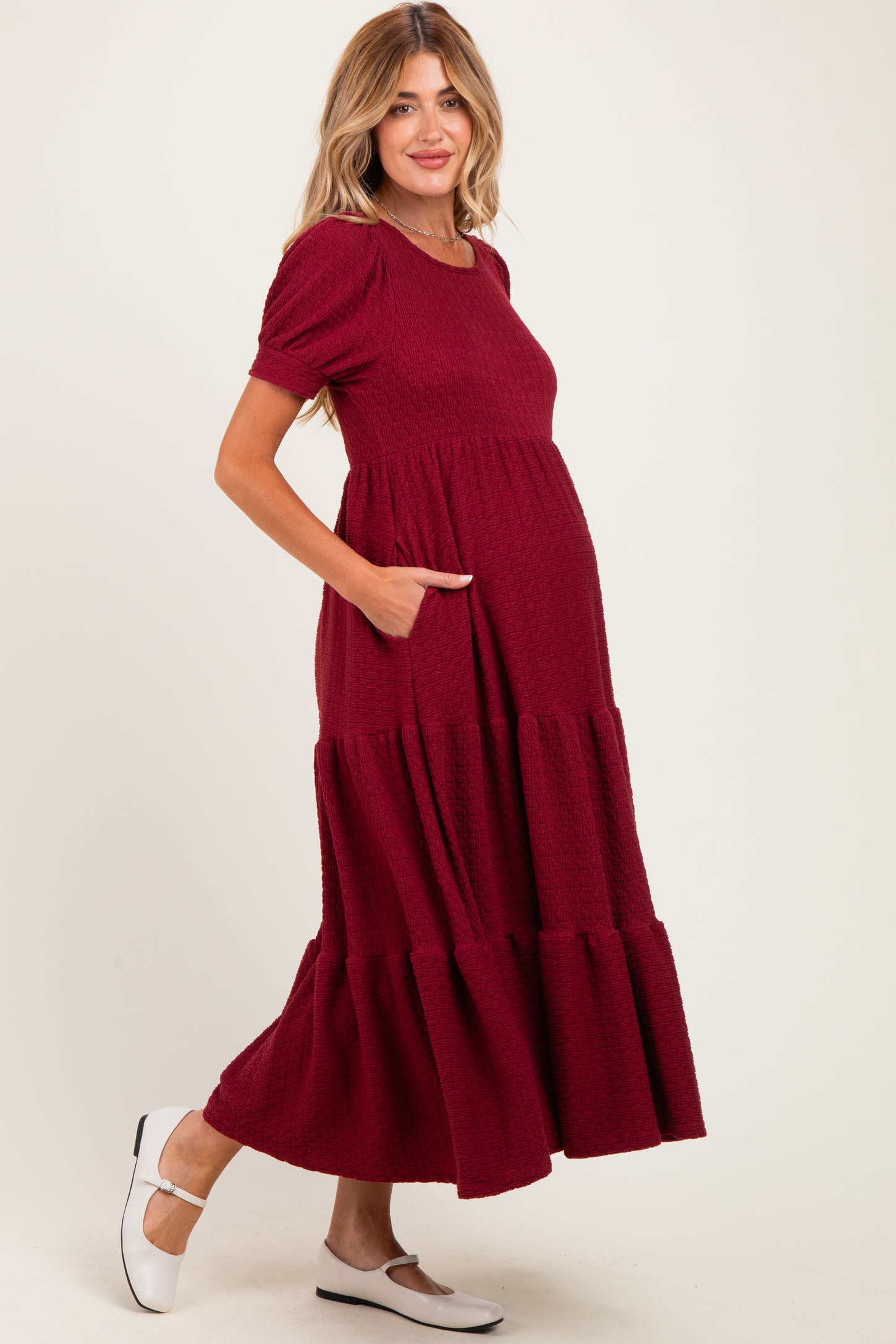 woman wearing a burgundy maternity maxi dress with puff sleeves and side pockets, showing her baby bump