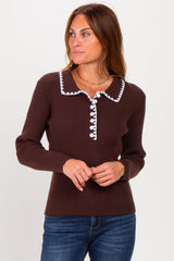 woman wearing a brown ribbed sweater with white pom-pom trim and a collared neckline, standing and looking to the side