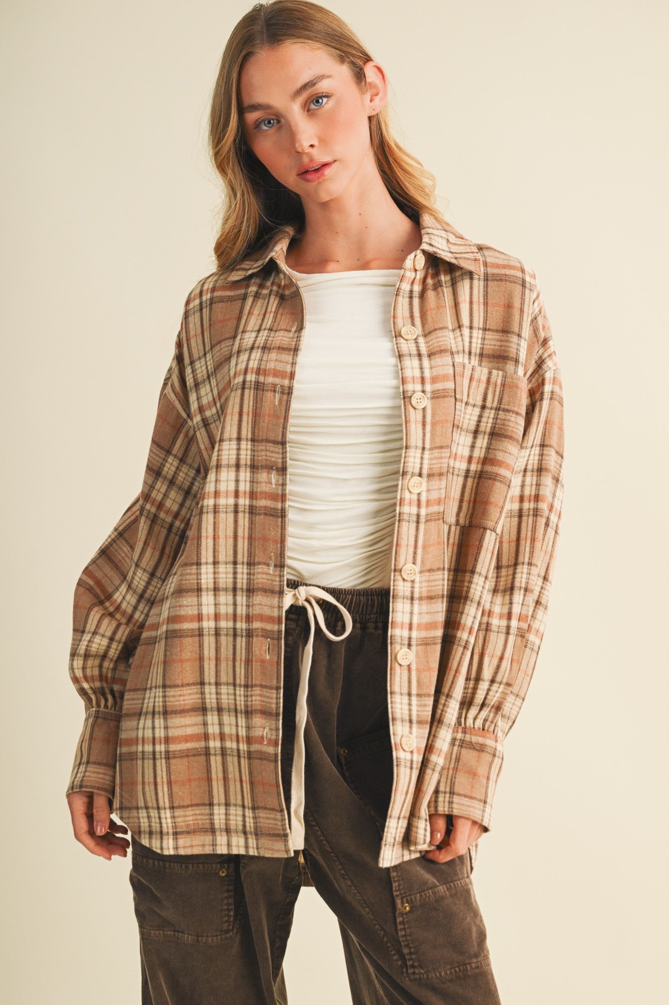 woman wearing a brown plaid oversized shirt over a white fitted top, styled with dark brown pants