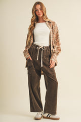woman wearing a white fitted top, brown wide-leg corduroy pants, and an oversized plaid shirt, standing against a light background