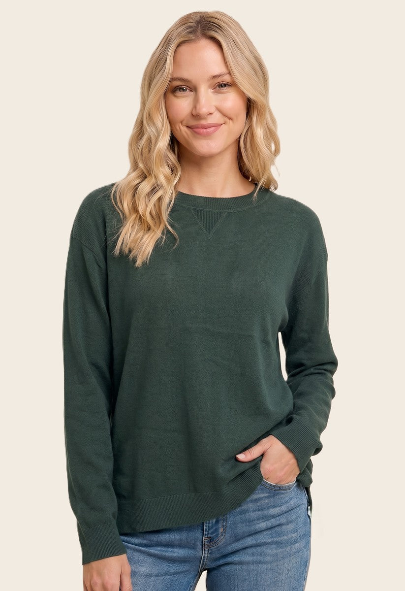 woman wearing a dark green long sleeve sweater and blue jeans, smiling with one hand in her pocket