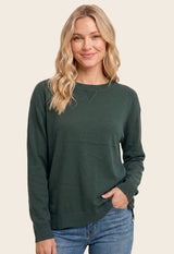 woman wearing a dark green long sleeve sweater and blue jeans, smiling with one hand in her pocket