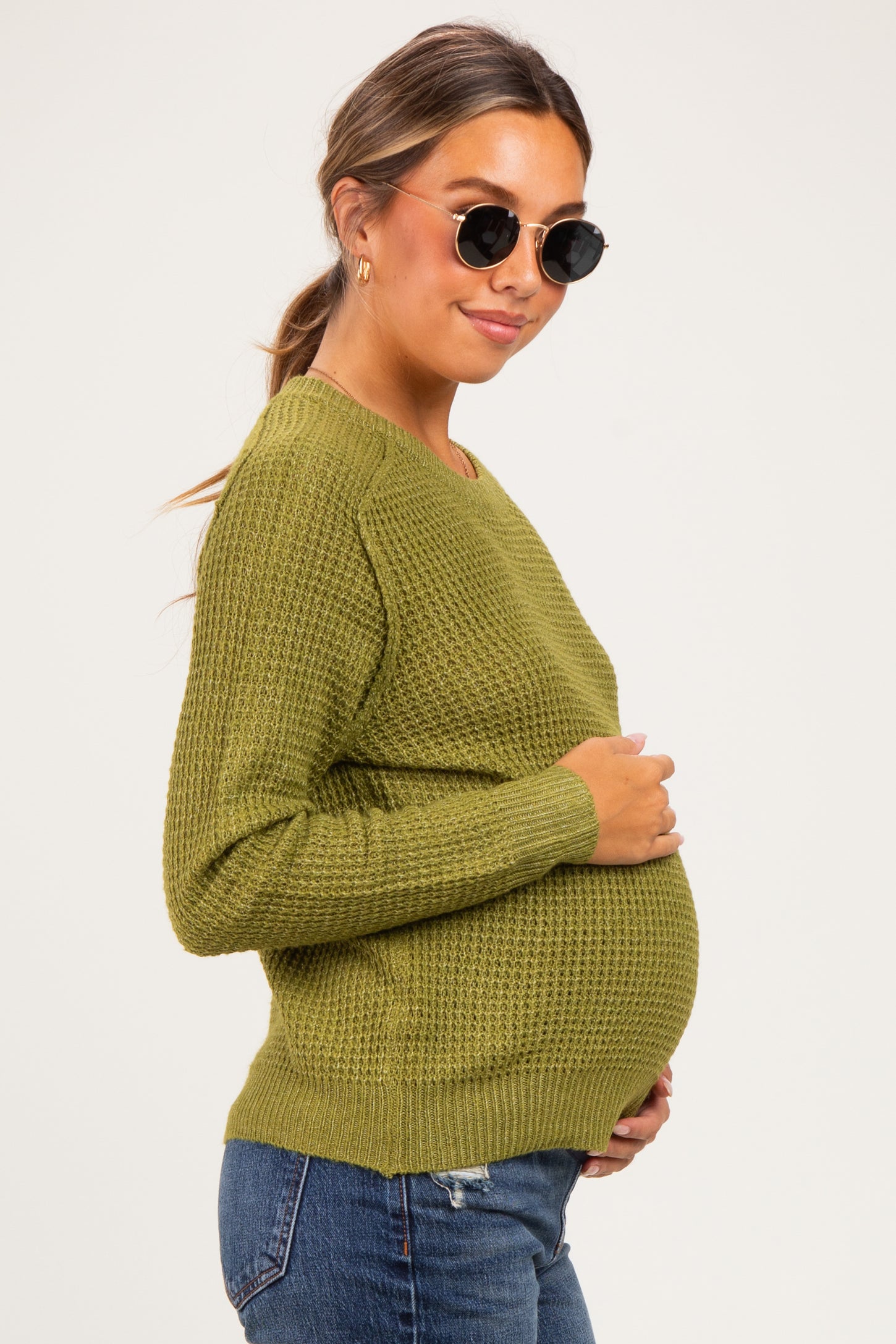 woman wearing an olive green textured maternity sweater and cradling her baby bump in a side view