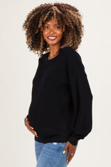 woman wearing a black maternity sweater, smiling and holding her baby bump, paired with blue jeans
