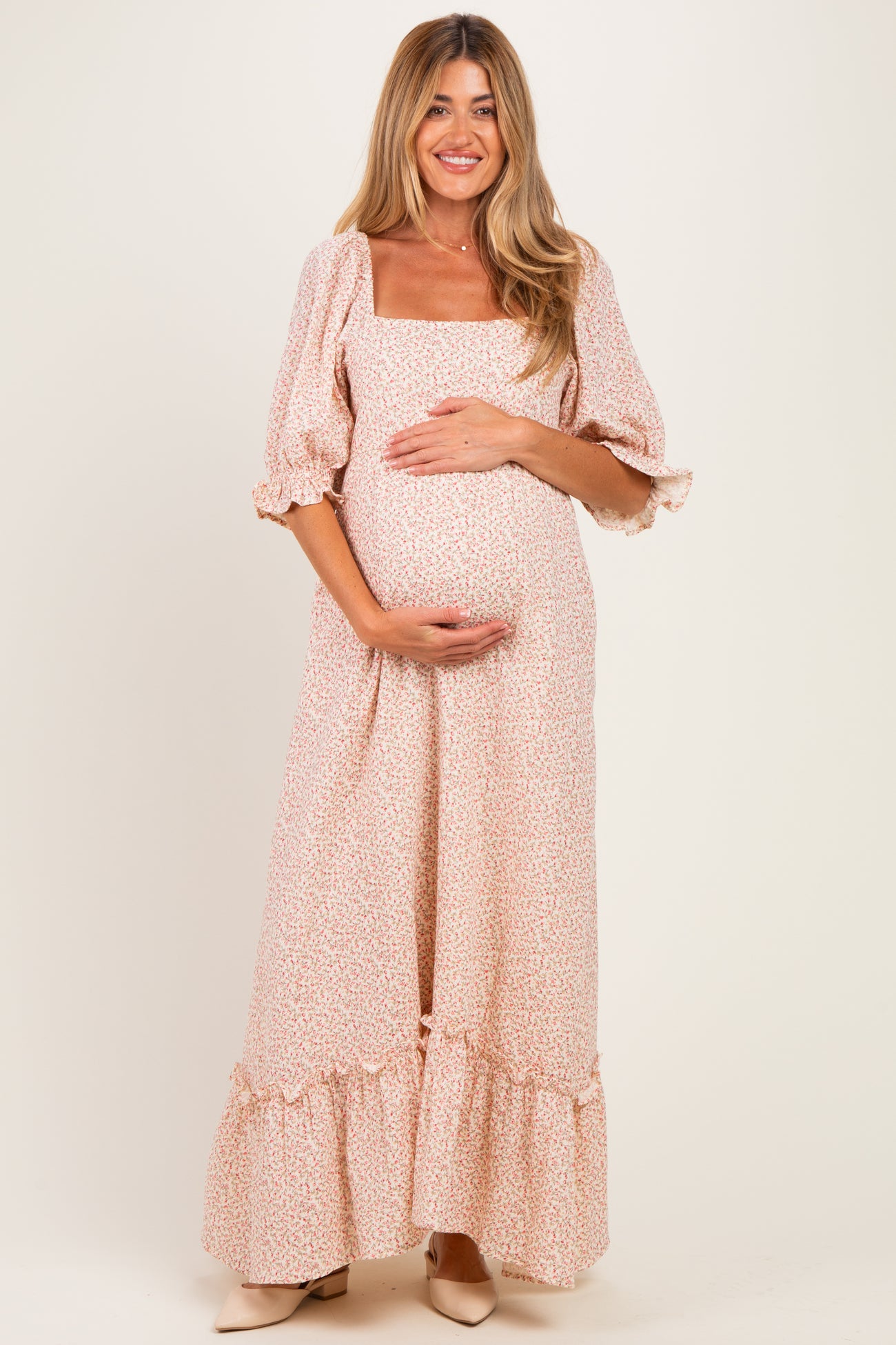woman wearing a blush floral maternity maxi dress with puff sleeves and ruffled hem, holding her baby bump
