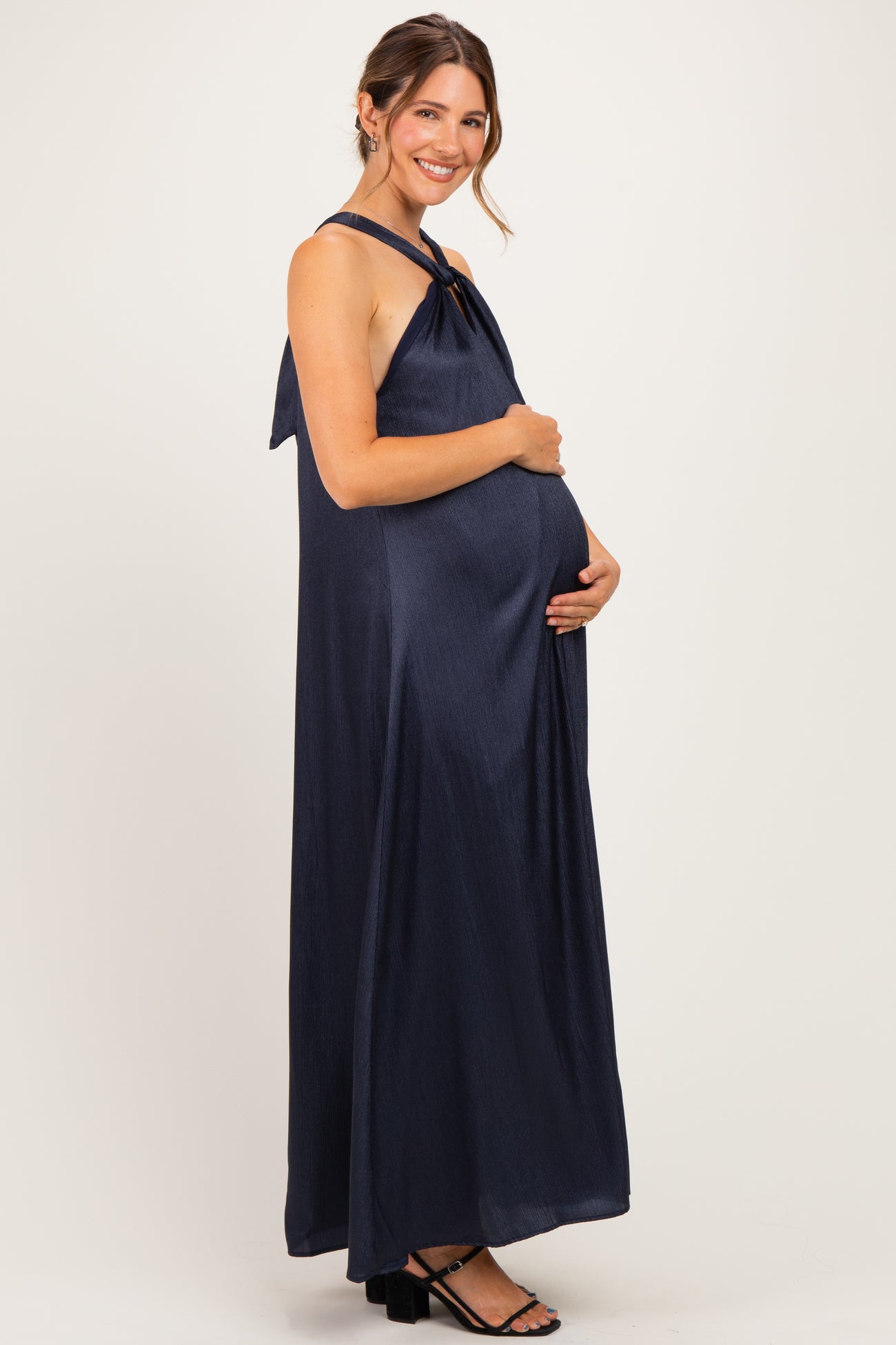 woman wearing a navy blue sleeveless maternity maxi dress with a tie back and holding her baby bump