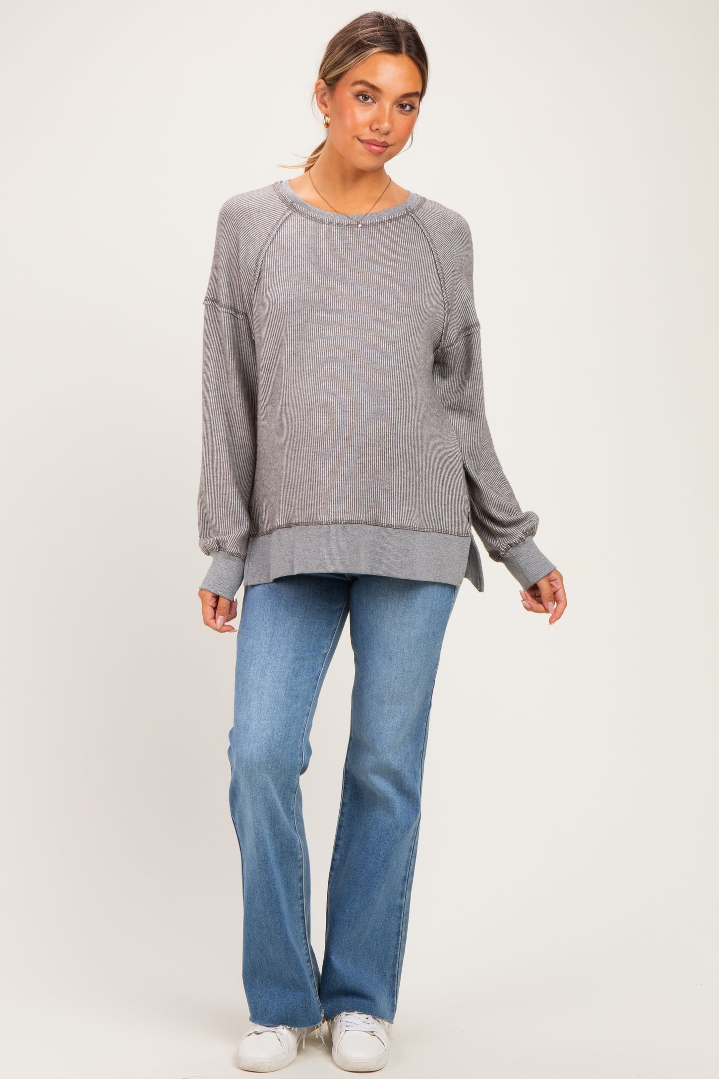 woman wearing a gray ribbed maternity sweatshirt with side slits, paired with light blue flared jeans and white sneakers