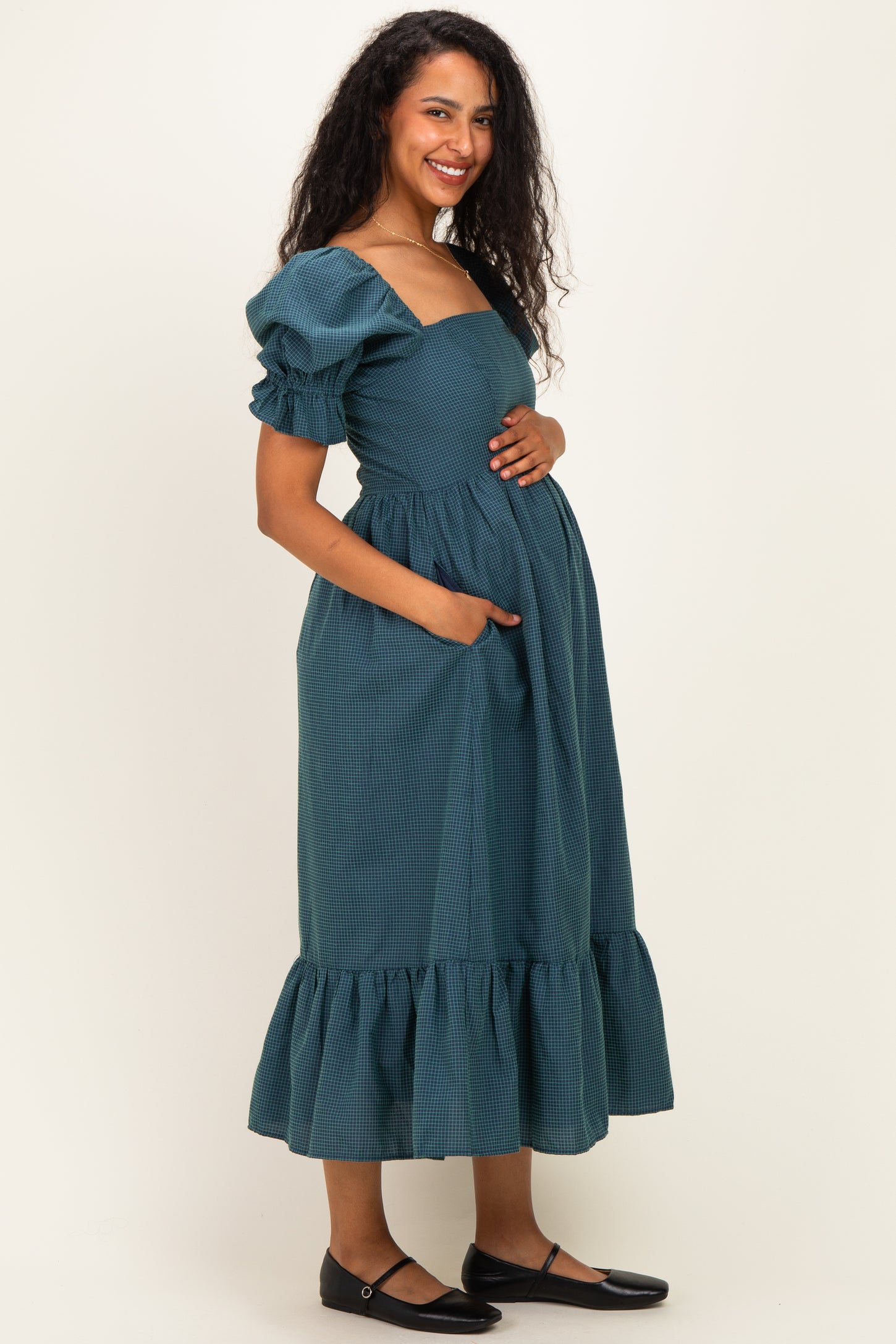 woman wearing a dark teal maternity dress with puff sleeves and ruffle hem, holding her baby bump