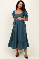 woman wearing a green maternity dress with puff sleeves and a tiered ruffle hem, holding her baby bump