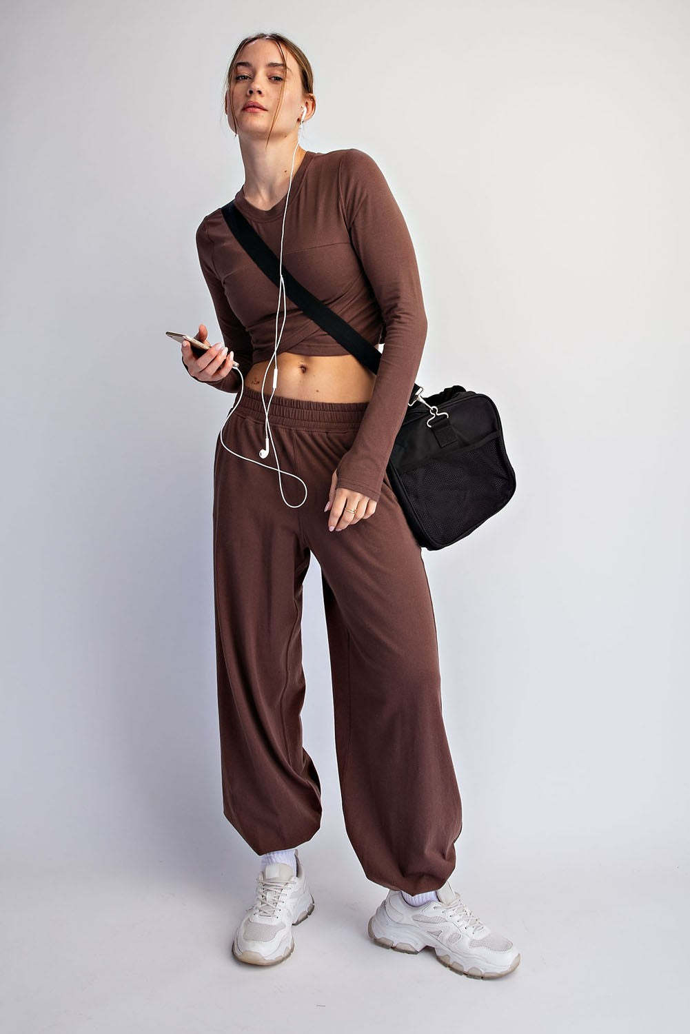 woman wearing a brown long sleeve crop top and matching wide-leg pants, holding a phone and wearing sneakers