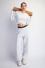 woman wearing a white cropped long sleeve top and light gray sweatpants, holding a yoga mat over her shoulders