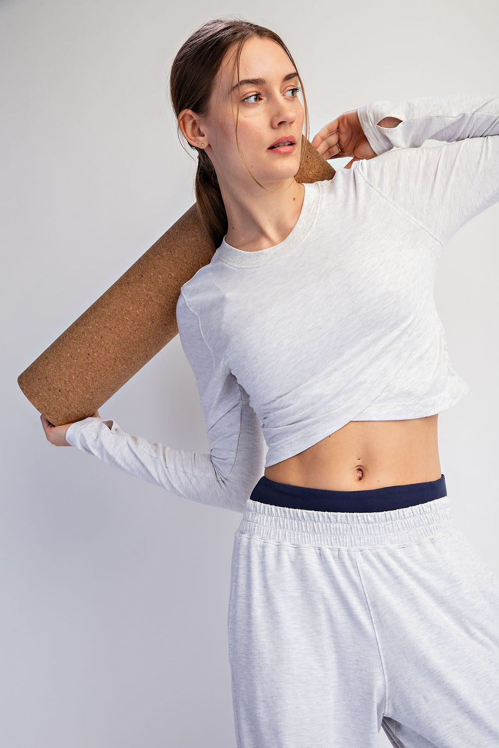 woman wearing a light gray long sleeve crop top and gray sweatpants, holding a yoga mat over her shoulder