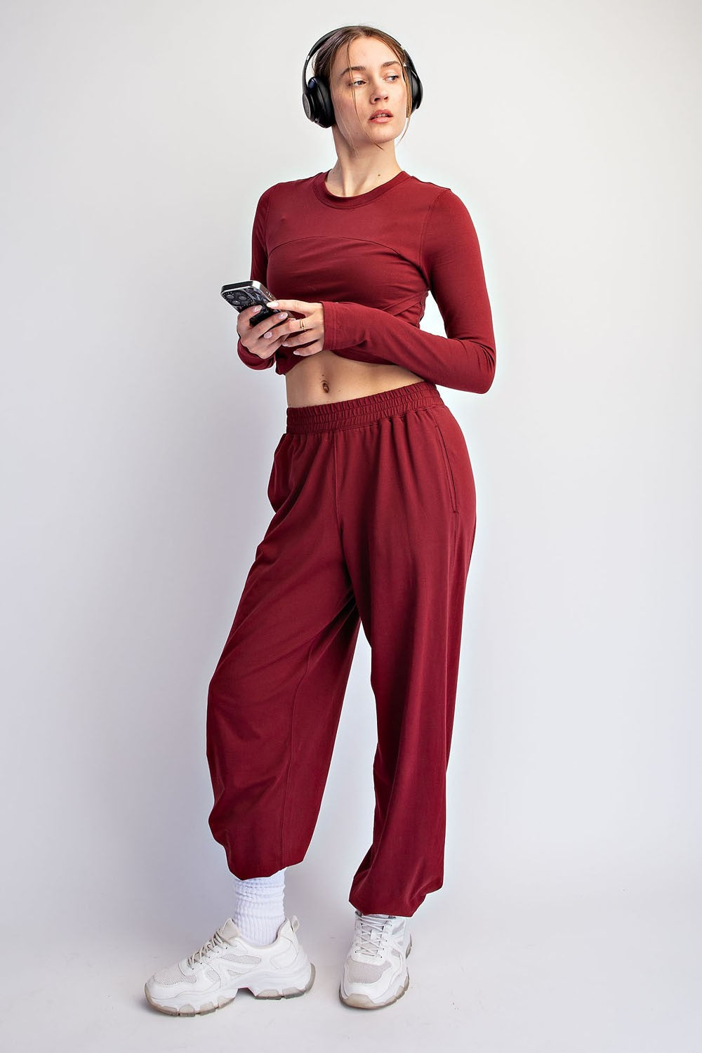 woman wearing a burgundy long sleeve crop top and matching jogger pants, holding a phone and wearing headphones