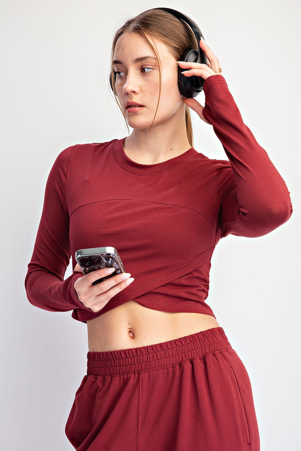 woman wearing a burgundy long sleeve crop top and matching joggers, holding a phone and wearing headphones