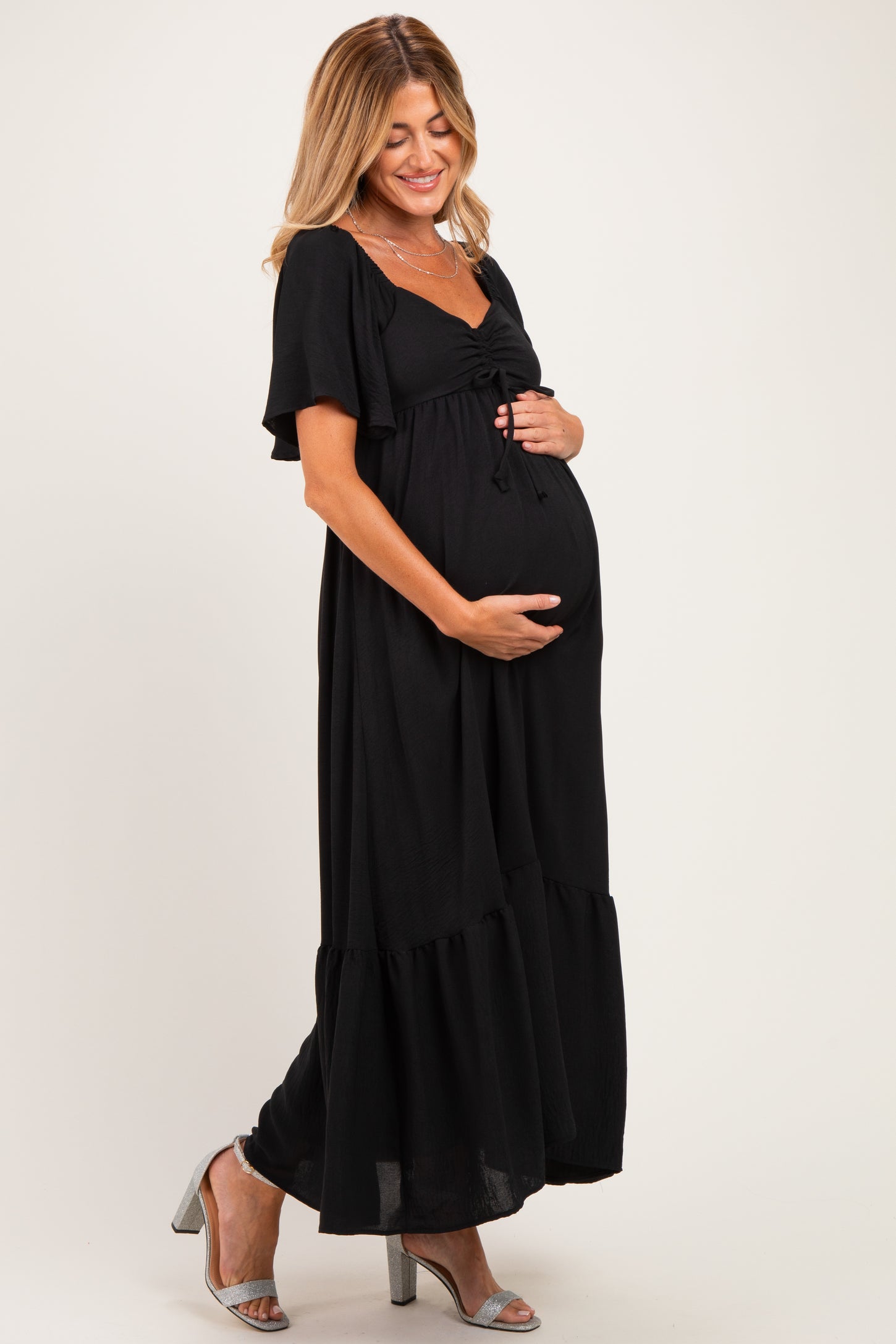 woman wearing a black maternity maxi dress with short sleeves and a tie detail, holding her baby bump