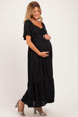 woman wearing a black maternity maxi dress with short sleeves and a tie detail, holding her baby bump