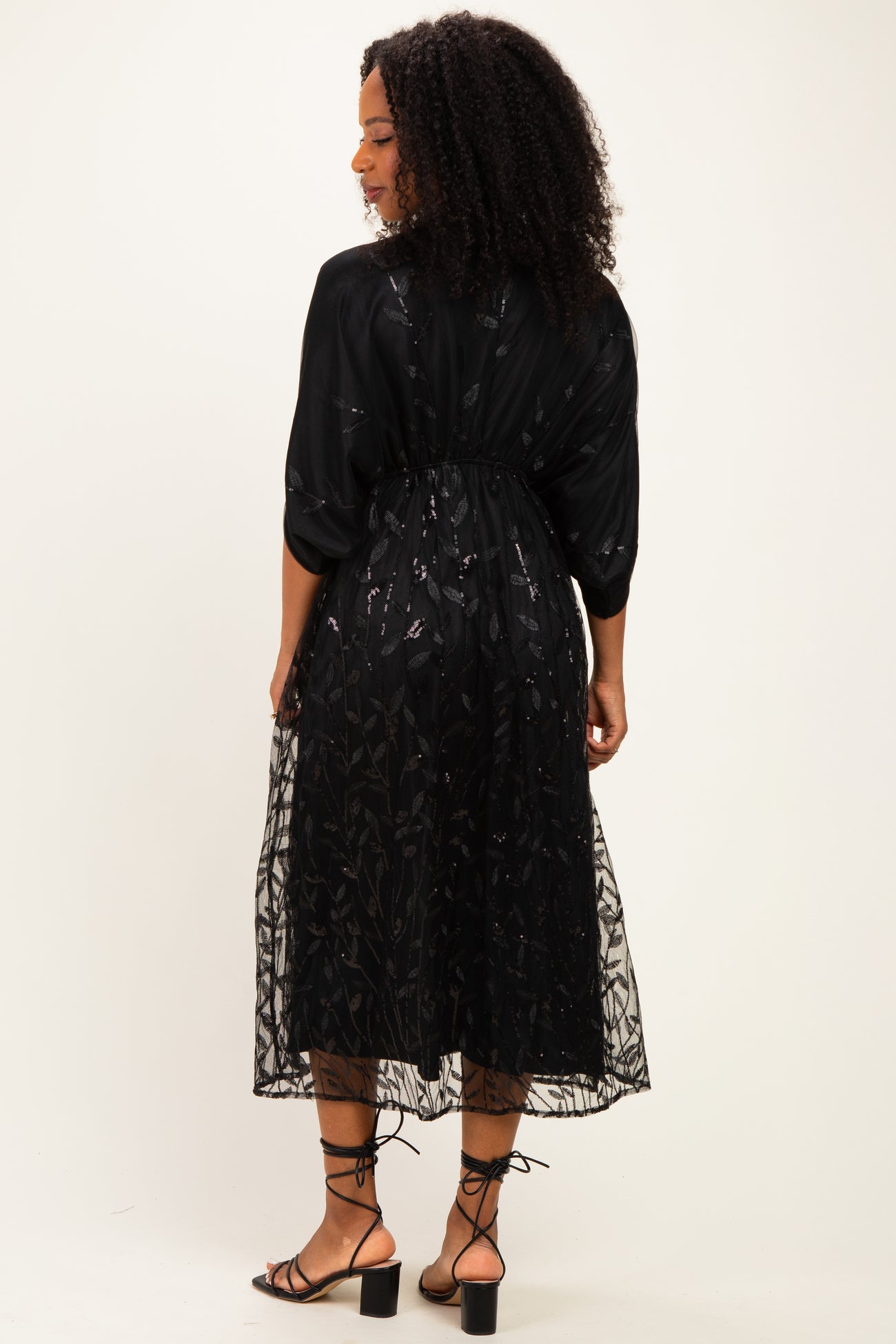 woman wearing a black sheer midi dress with floral embroidery and a cinched waist, showing the back view