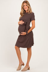 woman wearing a brown short sleeve maternity dress with a tie detail, gently cradling her baby bump