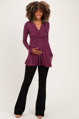 woman wearing a plum long sleeve maternity top with a v-neck and holding her baby bump, paired with black flared pants