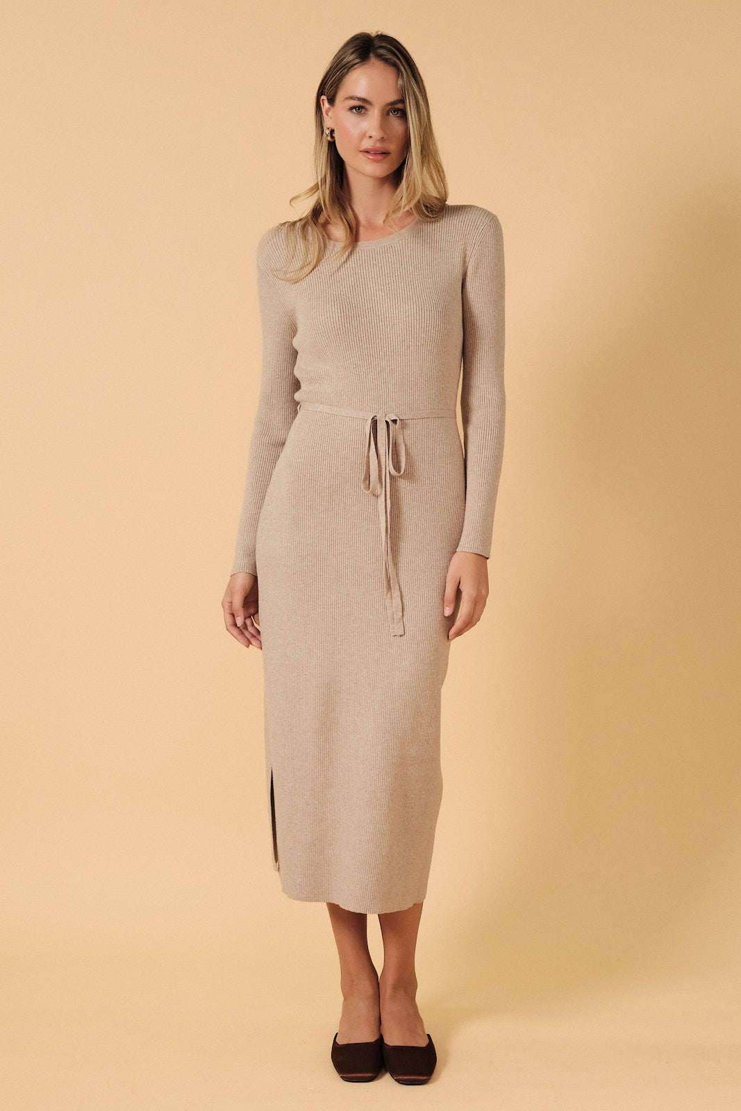 woman wearing a beige ribbed midi dress with long sleeves and a tie waist detail, standing against a light background