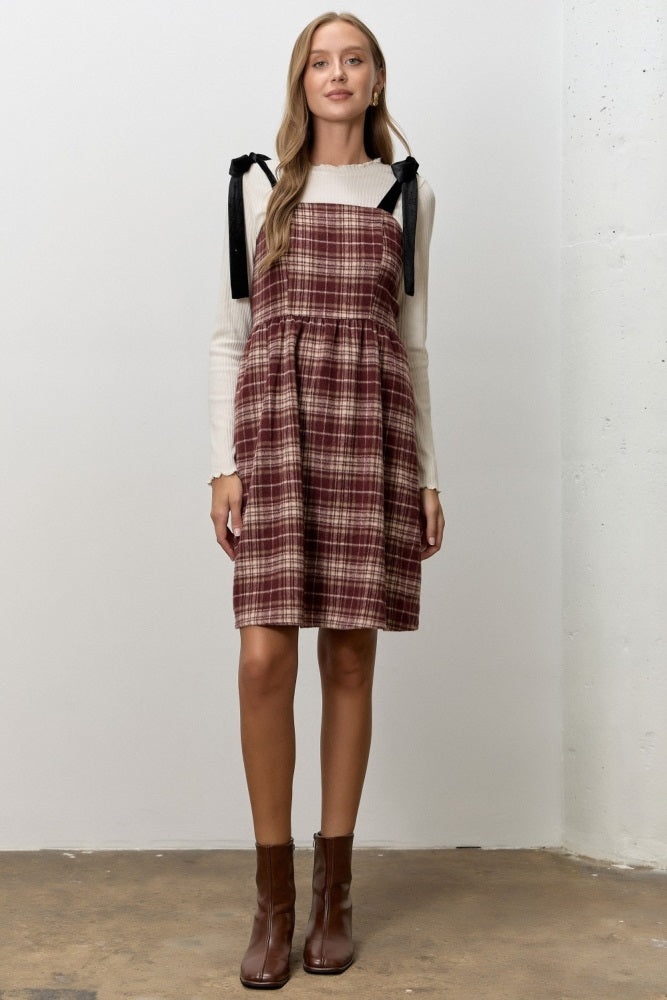woman wearing a burgundy plaid dress with black tie straps and a cream long sleeve top, standing on a concrete floor