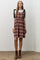 woman wearing a burgundy plaid dress with black tie straps and a cream long sleeve top, standing on a concrete floor