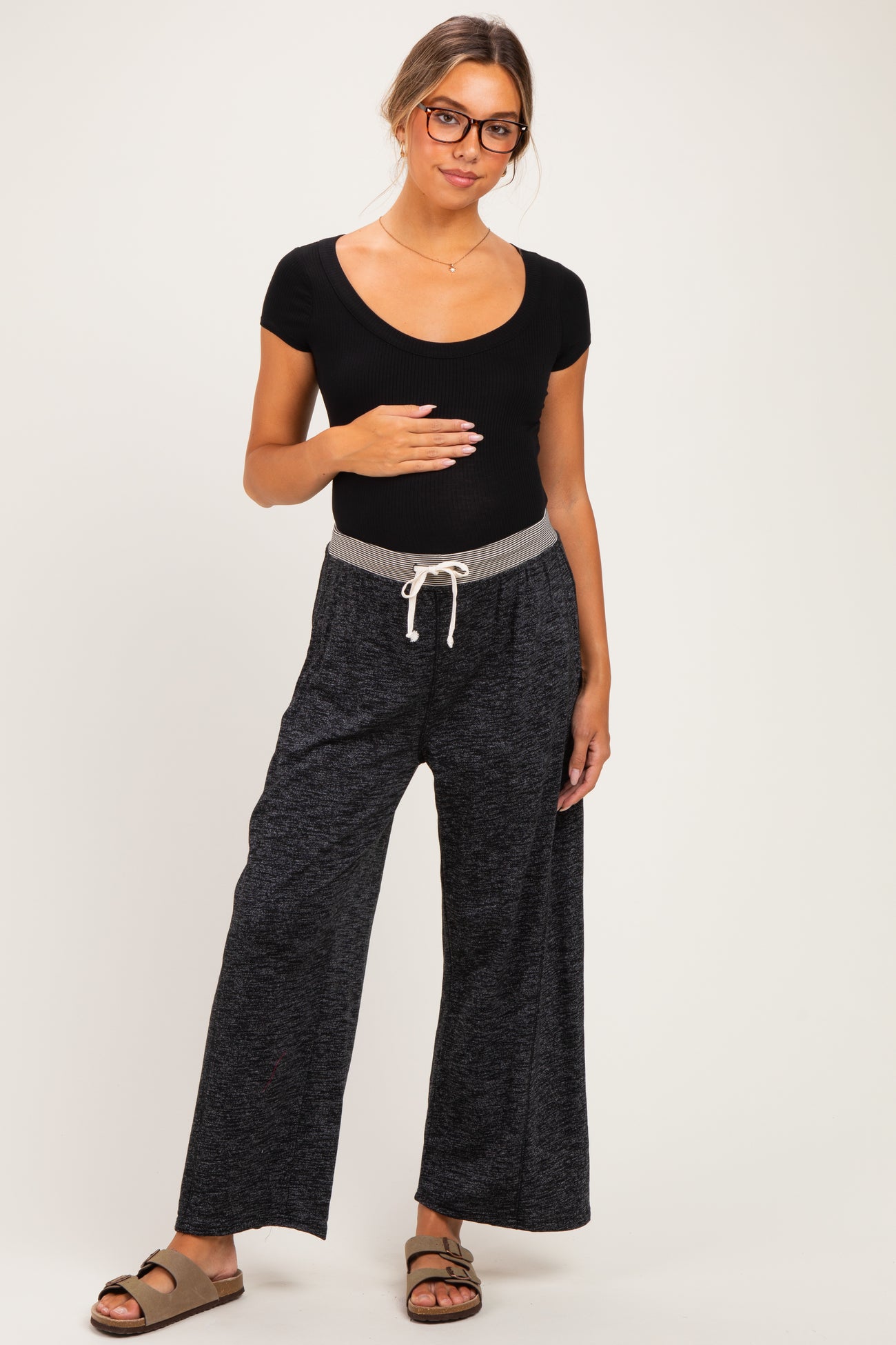 woman wearing a black ribbed maternity top and gray wide-leg maternity pants, holding her baby bump