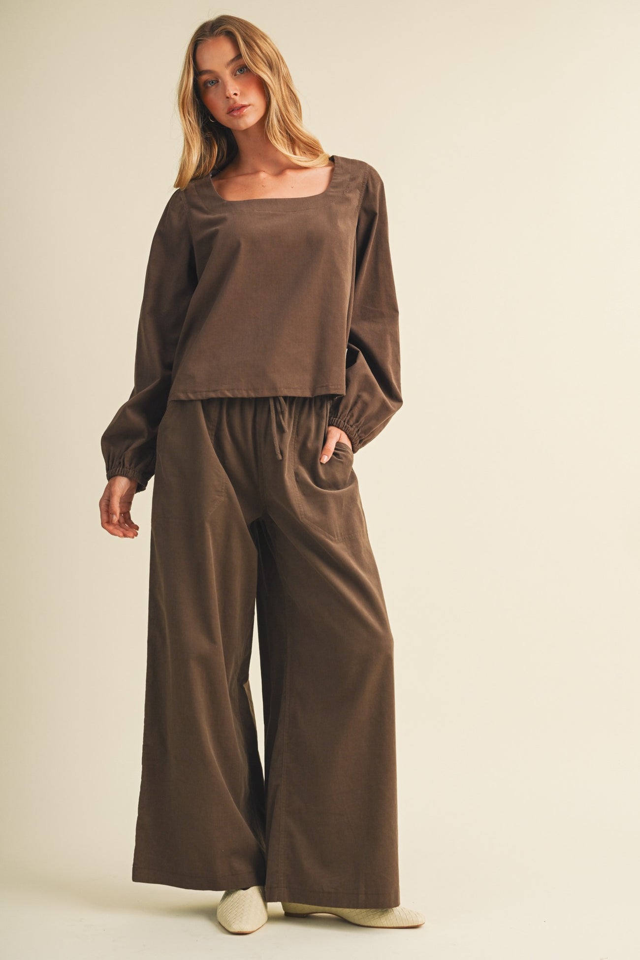 woman wearing a brown long sleeve top and matching wide-leg pants, posing with hands in pockets