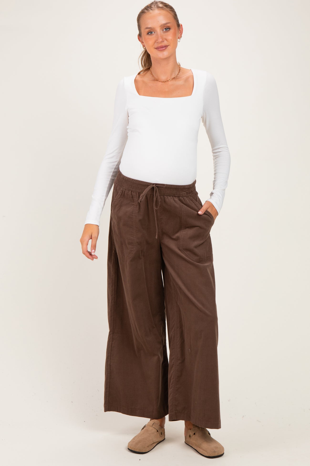 woman wearing a white long sleeve maternity top and brown wide-leg maternity pants, showing her baby bump