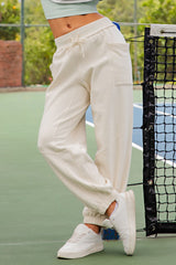 woman wearing cream-colored sweatpants with an elastic waistband and pockets, standing beside a tennis net