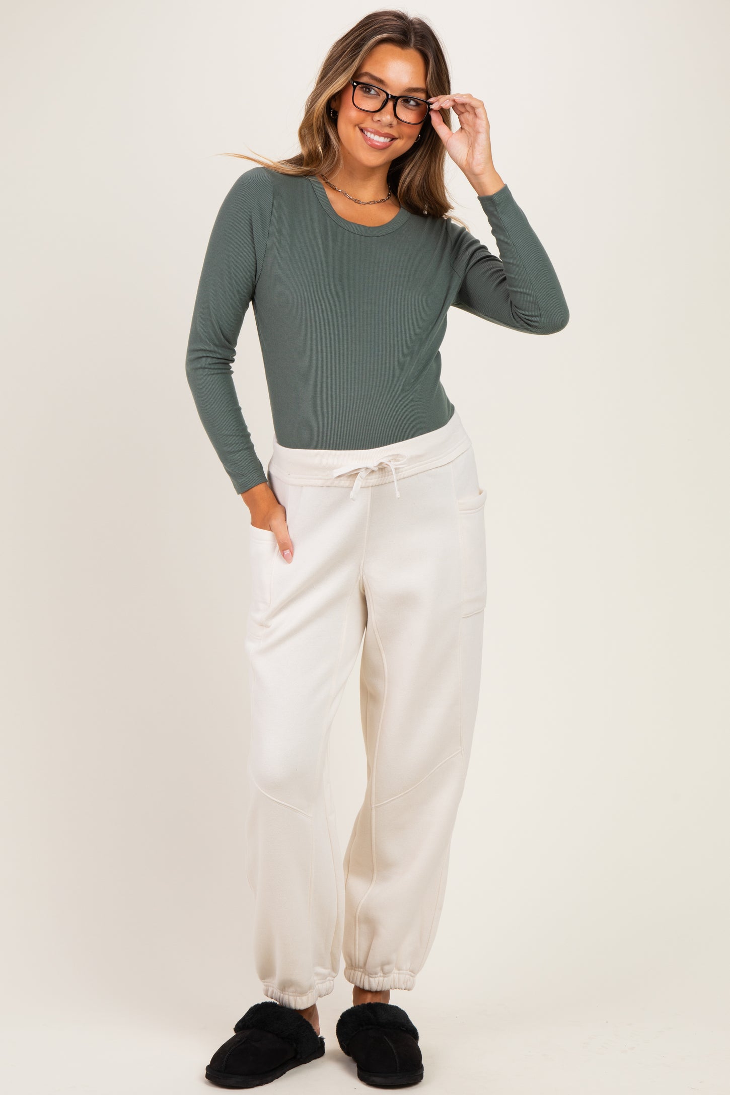 woman wearing a dark green long sleeve maternity top and light beige maternity joggers, smiling with glasses