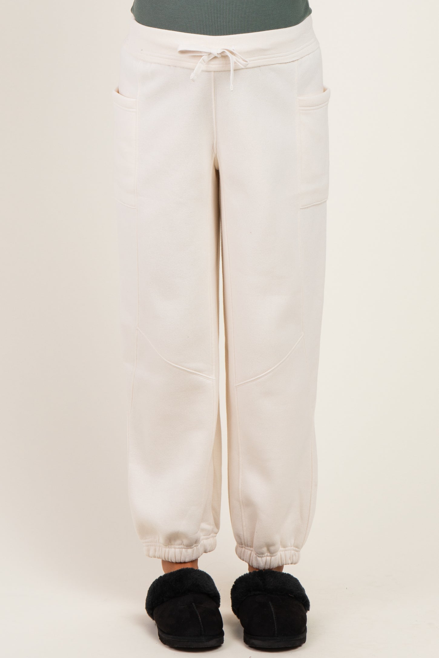woman wearing cream-colored maternity sweatpants with pockets and elastic cuffs, standing in plush slippers