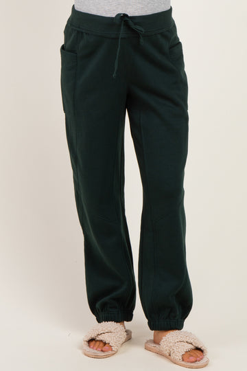 woman wearing dark green maternity jogger pants with an elastic waistband and pockets, styled with fuzzy slippers