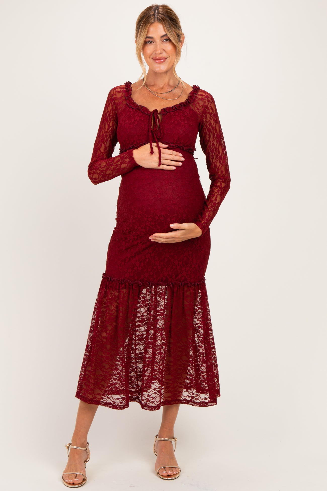 woman wearing a burgundy lace maternity dress with long sleeves and a tiered skirt, cradling her baby bump