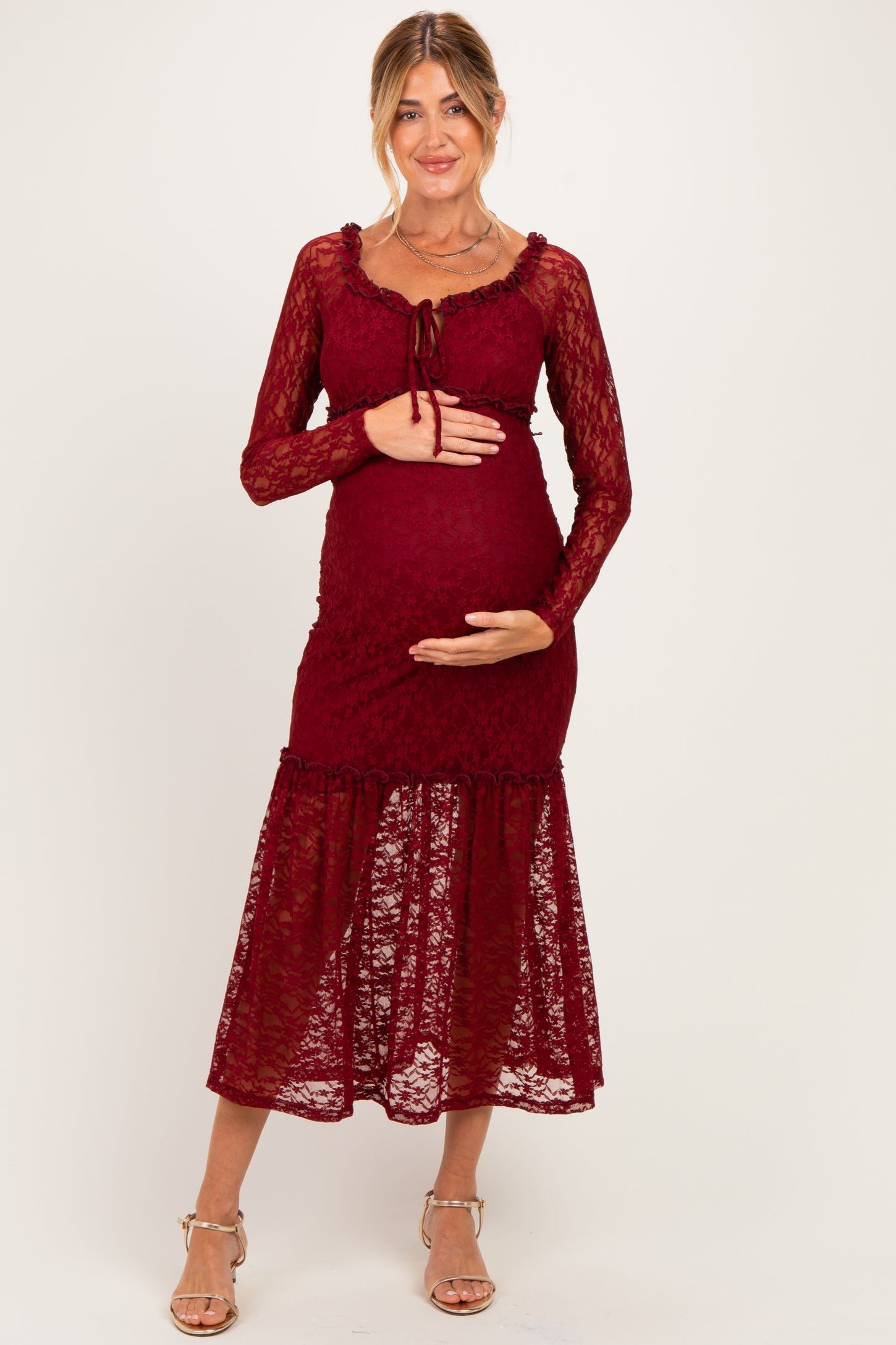 woman wearing a burgundy lace maternity dress with long sleeves and a tiered skirt, cradling her baby bump