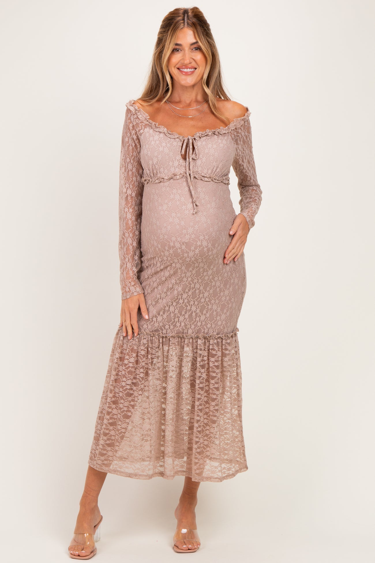 woman wearing a light brown lace maternity dress with long sleeves and a flowing hem, gently cradling her baby bump