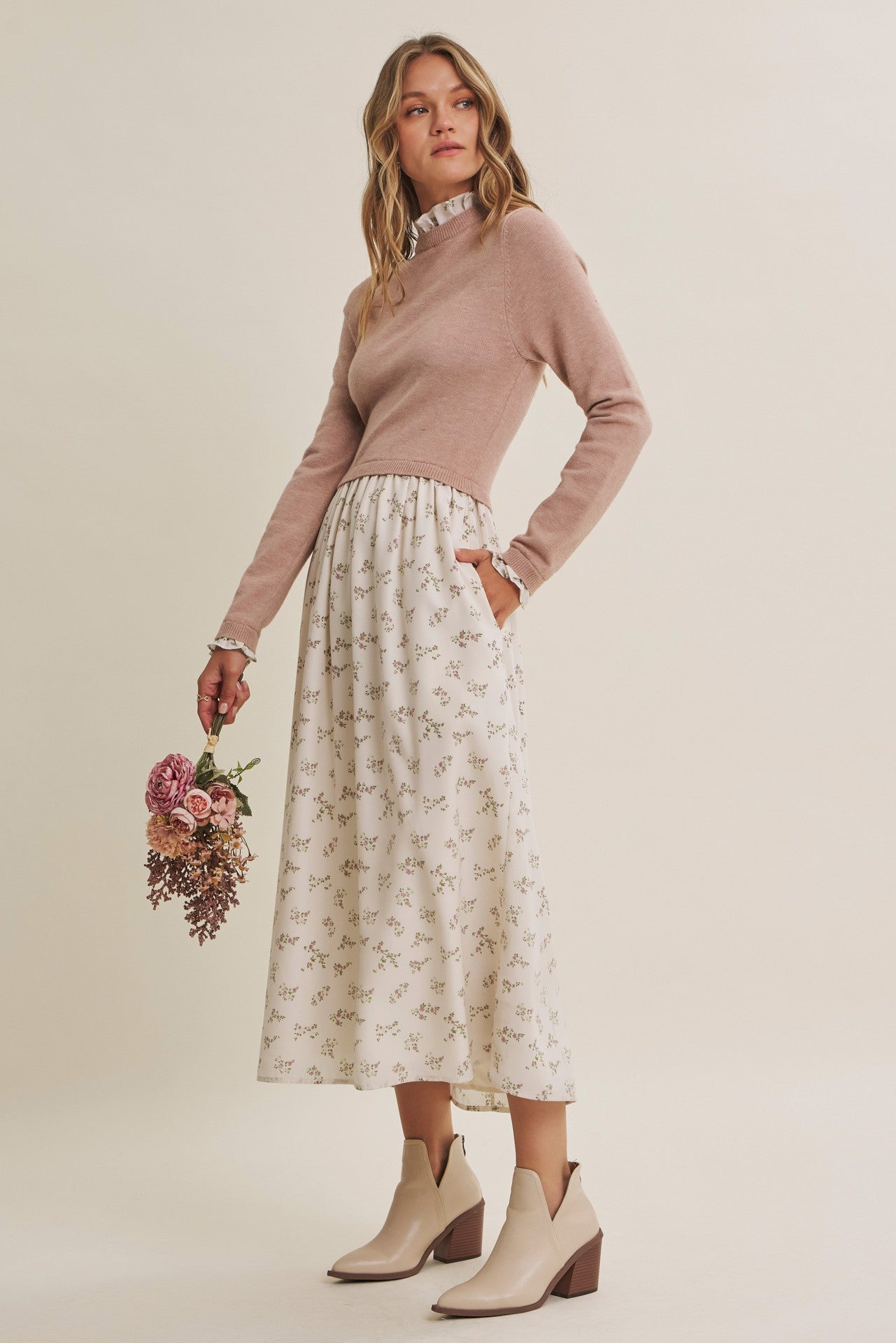 woman wearing a light brown sweater and a cream floral midi skirt, holding a bouquet of flowers